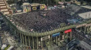 Aerial view of a crowded stadium filled with people attending a concert. The stage is adorned with large, colorful decorations, and the surrounding area includes visible infrastructure and banners.